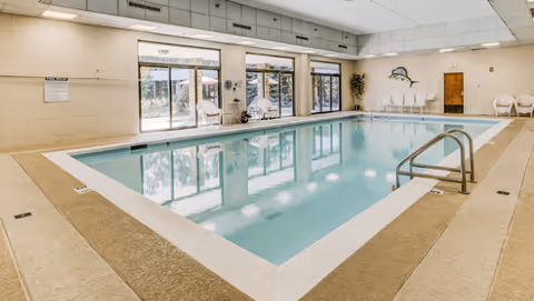 Indoor swimming pool with clear blue water, surrounded by beige tiled flooring and white walls. Large windows and glass doors line one side, allowing natural light to enter. Several white chairs are placed along the walls, and a metal handrail leads into the pool. A decorative fish wall art is visible on the far wall.