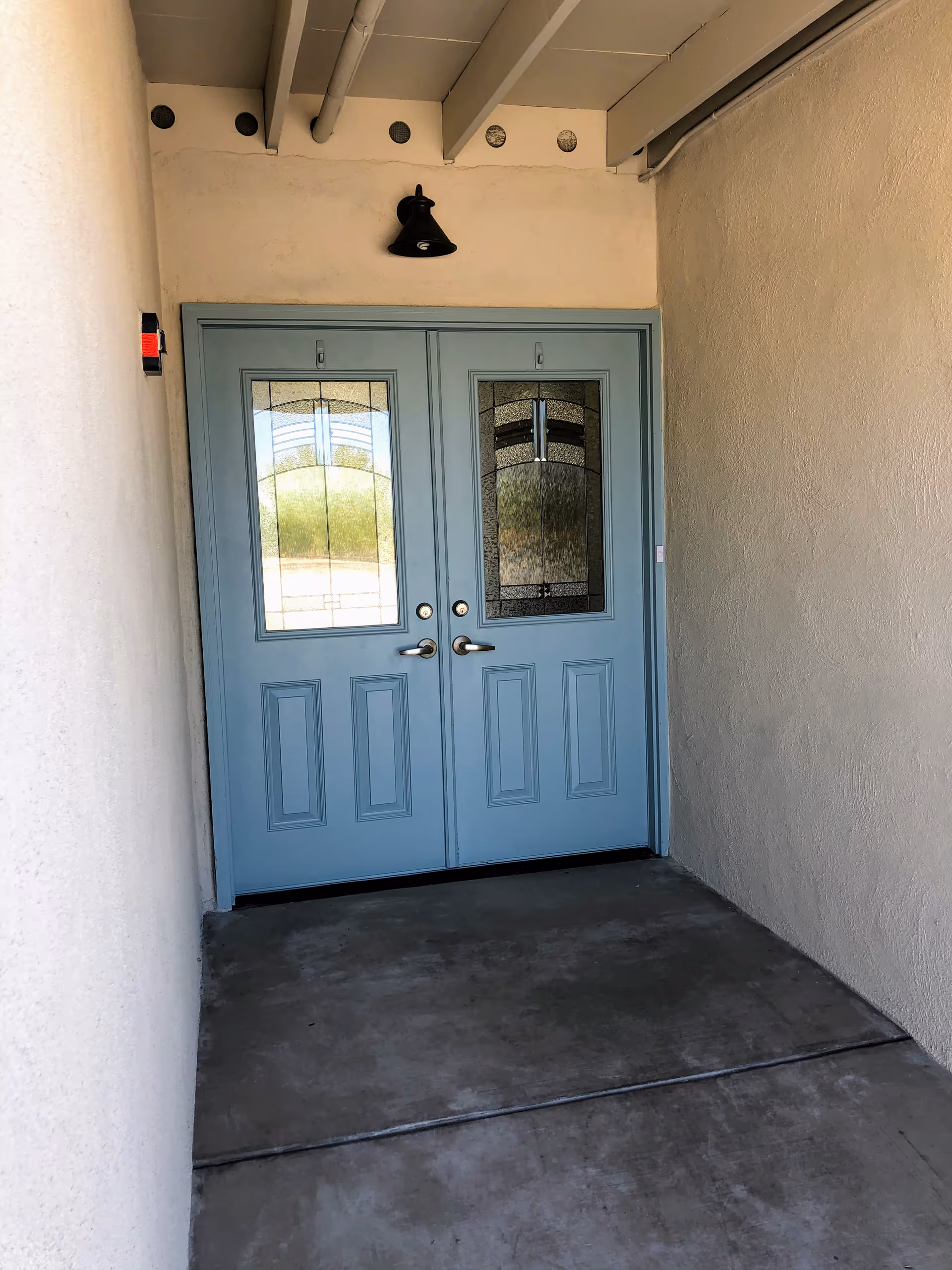A pair of light blue double doors with decorative glass panels set in a beige stucco wall under a covered entryway with a concrete floor and a black wall-mounted light fixture above the doors.