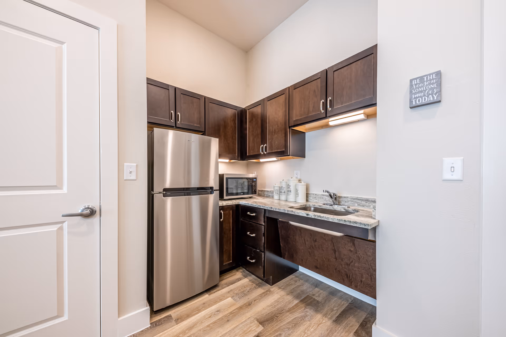 A small kitchen area with dark wood cabinets, a stainless steel refrigerator, a microwave on the countertop, a sink, and decorative containers. The floor has wood-style flooring and the walls are painted light gray. A small wall sign reads 'Be the reason someone smiles today.'