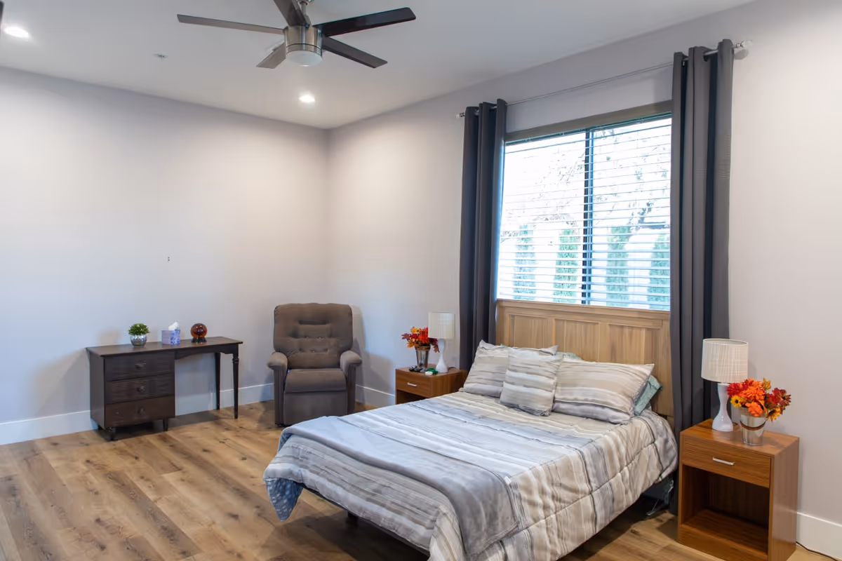 A bedroom with a wooden bed frame and a neatly made bed with striped bedding and multiple pillows. There are two wooden nightstands on either side of the bed, each with a lamp and a small flower arrangement. A large window with blinds and dark curtains is behind the bed. To the left, there is a dark wooden desk with a small plant, a tissue box, and a decorative item on top. Next to the desk is a cushioned armchair. The room has light-colored walls, wooden flooring, and a ceiling fan with lights.