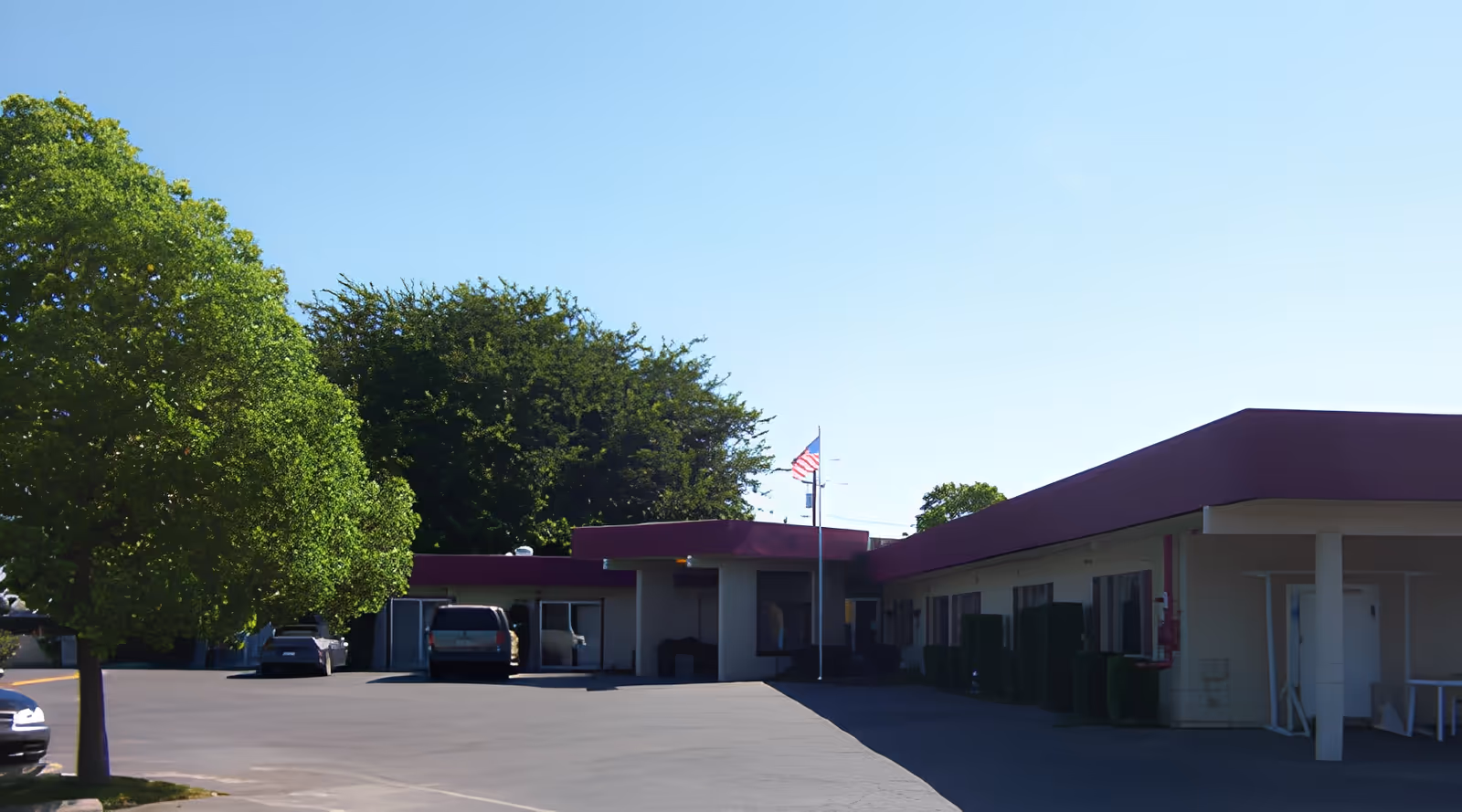 Exterior view of a single-story building with a flat roof and maroon trim, surrounded by trees and a parking area with several parked cars. An American flag is flying on a flagpole near the entrance.