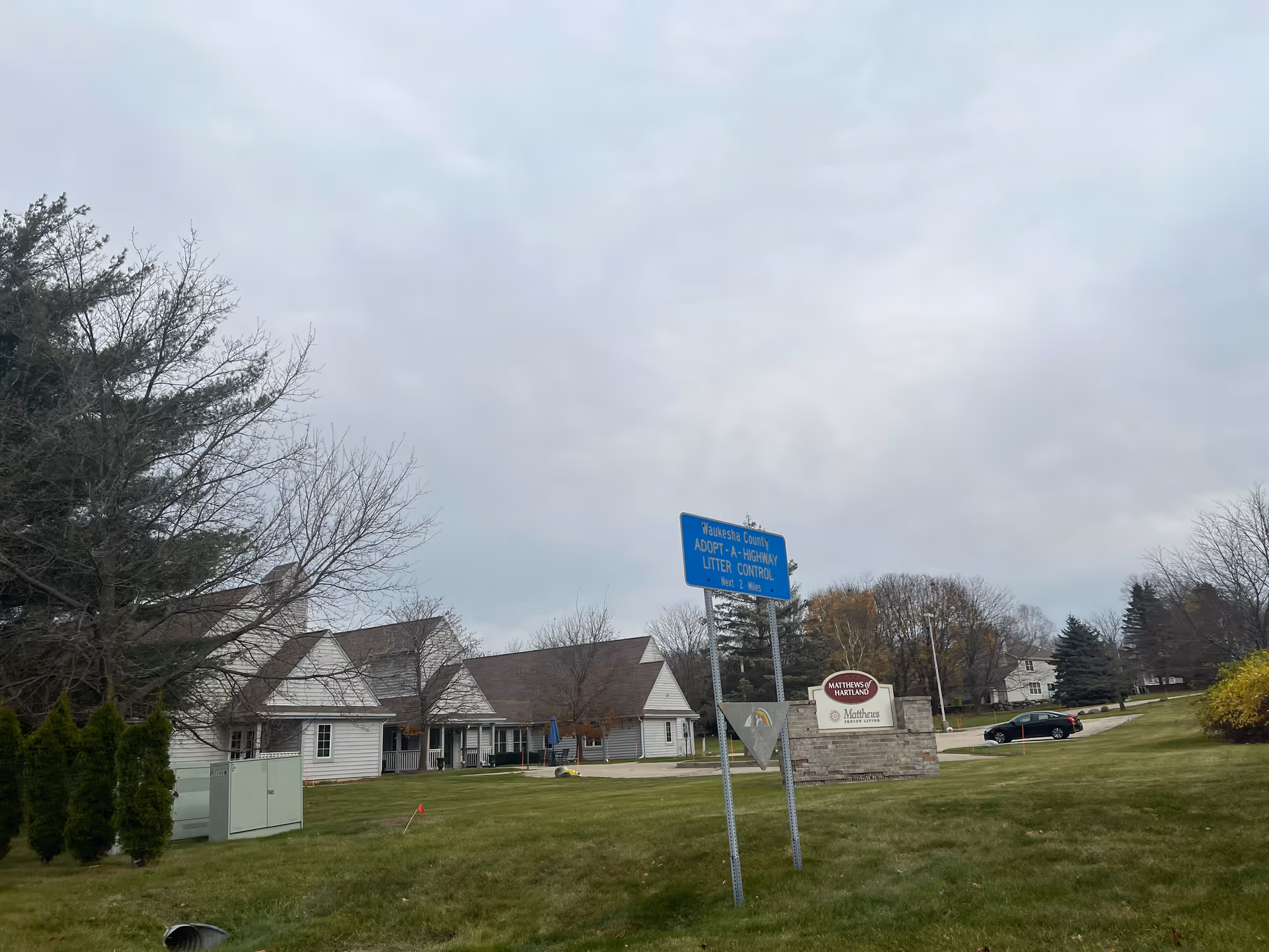 Exterior view of Matthews Of Hartland senior living facility with a large grassy area in the foreground, several trees, and a cloudy sky. There is a blue road sign indicating Waukesha County Adopt-A-Highway Litter Control for the next 2 miles, and a facility sign with the Matthews Of Hartland logo near the parking area with a black car parked.