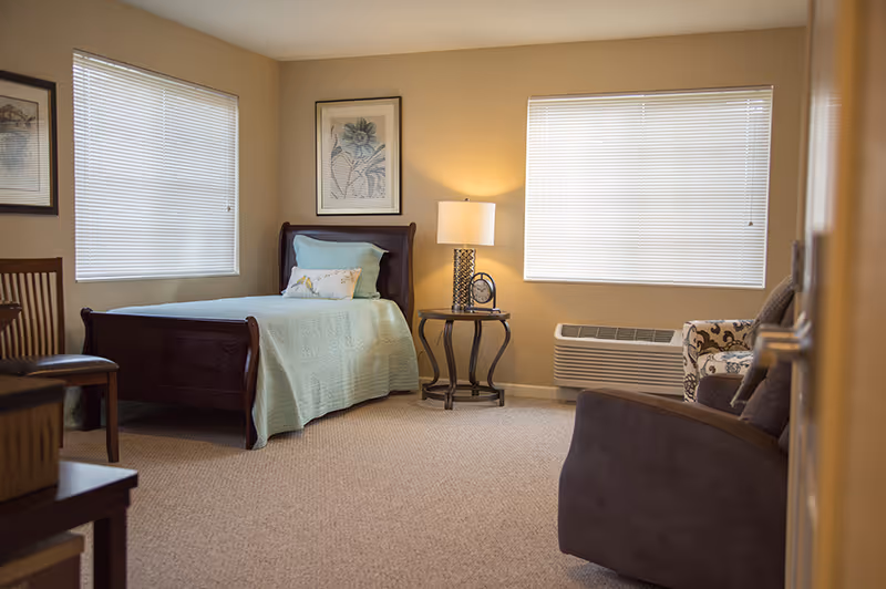 A cozy senior living bedroom with a single wooden bed covered in a light green quilt and pillows. The room has beige walls and carpet, two windows with closed blinds, a round side table with a lamp and a clock, a wooden chair, and two armchairs, one patterned and one solid-colored.