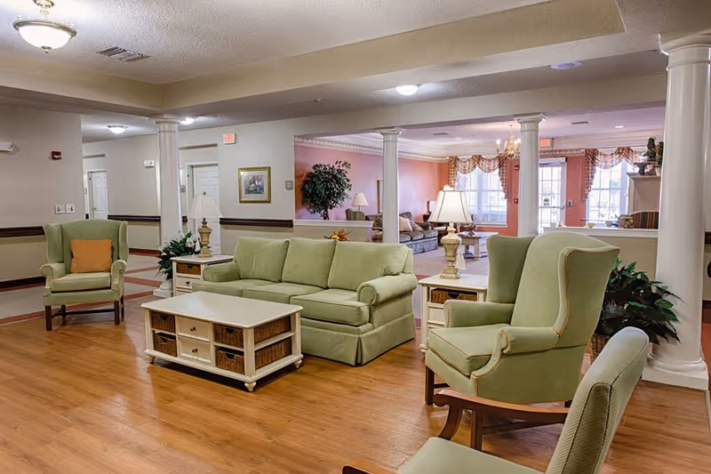 A spacious senior living facility common area with light green upholstered sofas and armchairs arranged around a white coffee table with wicker baskets. The room features wooden flooring, white columns, table lamps, and large windows with curtains allowing natural light to fill the space. There are plants and framed artwork on the walls, creating a warm and inviting atmosphere.