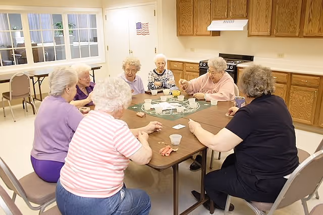 A group of elderly women sitting around a rectangular table playing a card game in a room with wooden cabinets and a stove in the background.