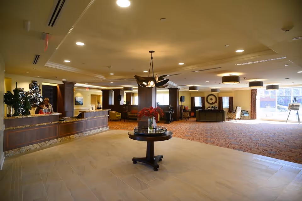 A spacious and well-lit reception area in a senior living facility with a wooden front desk on the left where two staff members are standing. In the center, there is a round table with a vase of red flowers. The room has a mix of tiled and carpeted flooring, with comfortable seating areas and large windows letting in natural light.