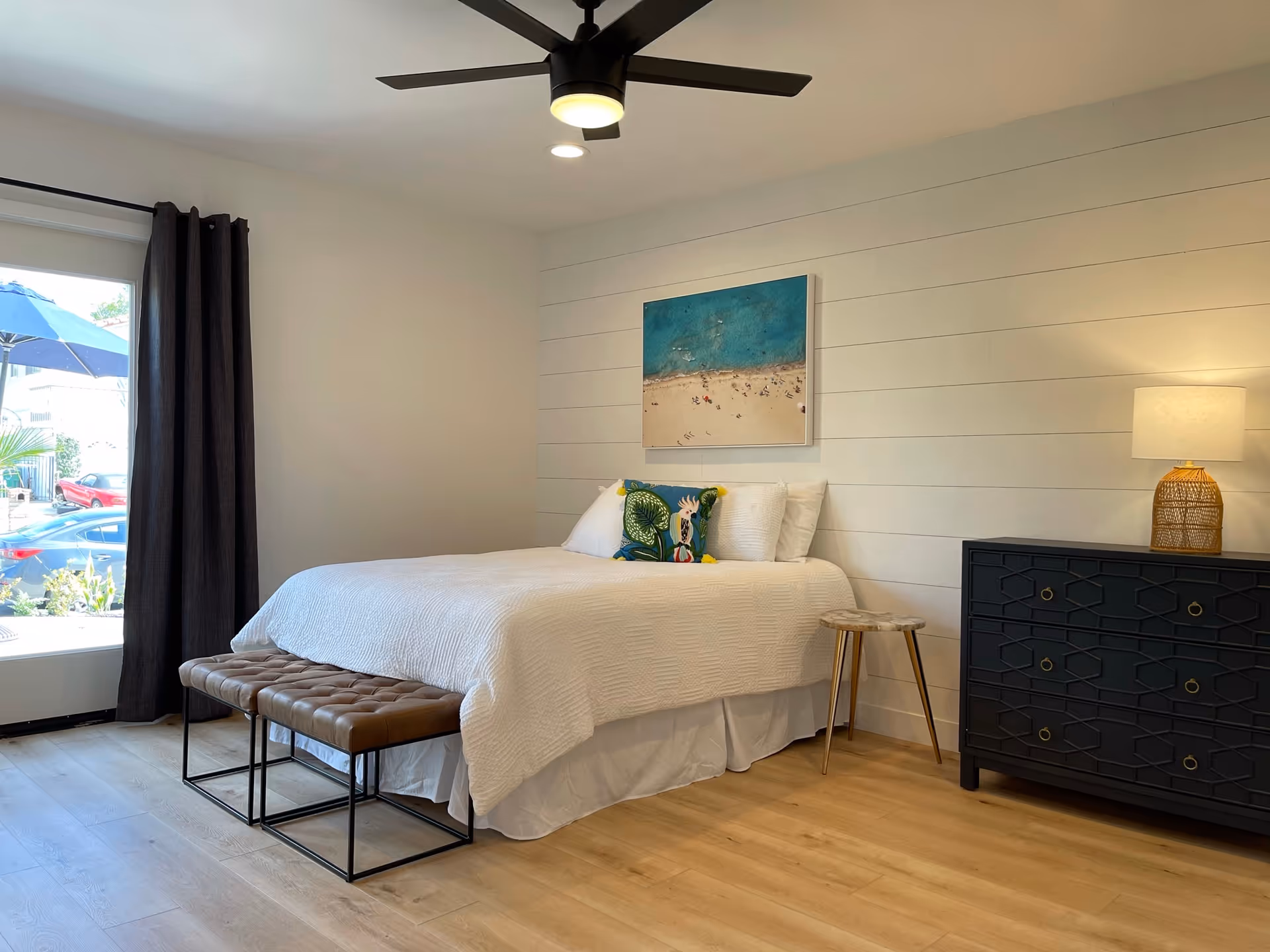 A bright bedroom with a white bedspread on a bed, two decorative pillows, a black dresser with a lamp, a small wooden side table, a ceiling fan with light, and a large window with dark curtains showing a view of parked cars and an outdoor umbrella.