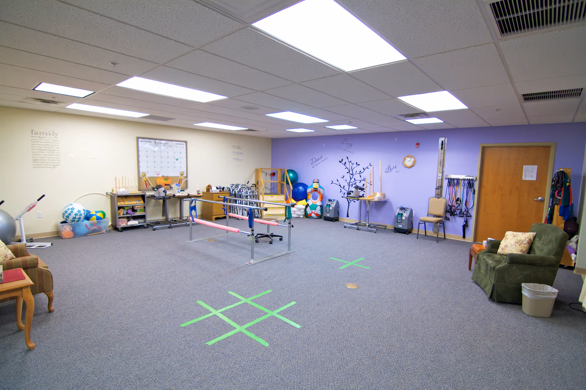 A spacious rehabilitation room with exercise equipment including parallel bars, therapy balls, walkers, and resistance bands. The walls are decorated with motivational words and a calendar. There are comfortable chairs and a small table along the sides of the room.