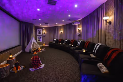 A cozy home theater room with a large projection screen on the left wall, a row of black reclining chairs with red and white blankets, dim wall sconces, and a ceiling with star-like purple lighting. There is a small white teepee tent with stuffed animals and decorative logs on the carpeted floor.