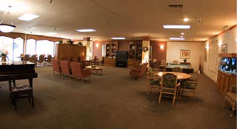 A spacious common area in a senior living facility featuring multiple seating arrangements including armchairs and tables with chairs. There is a piano on the left side, a large fish tank on the right, and a fireplace with a TV above it in the center background. The room is well-lit with ceiling lights and natural light from large windows on the left.
