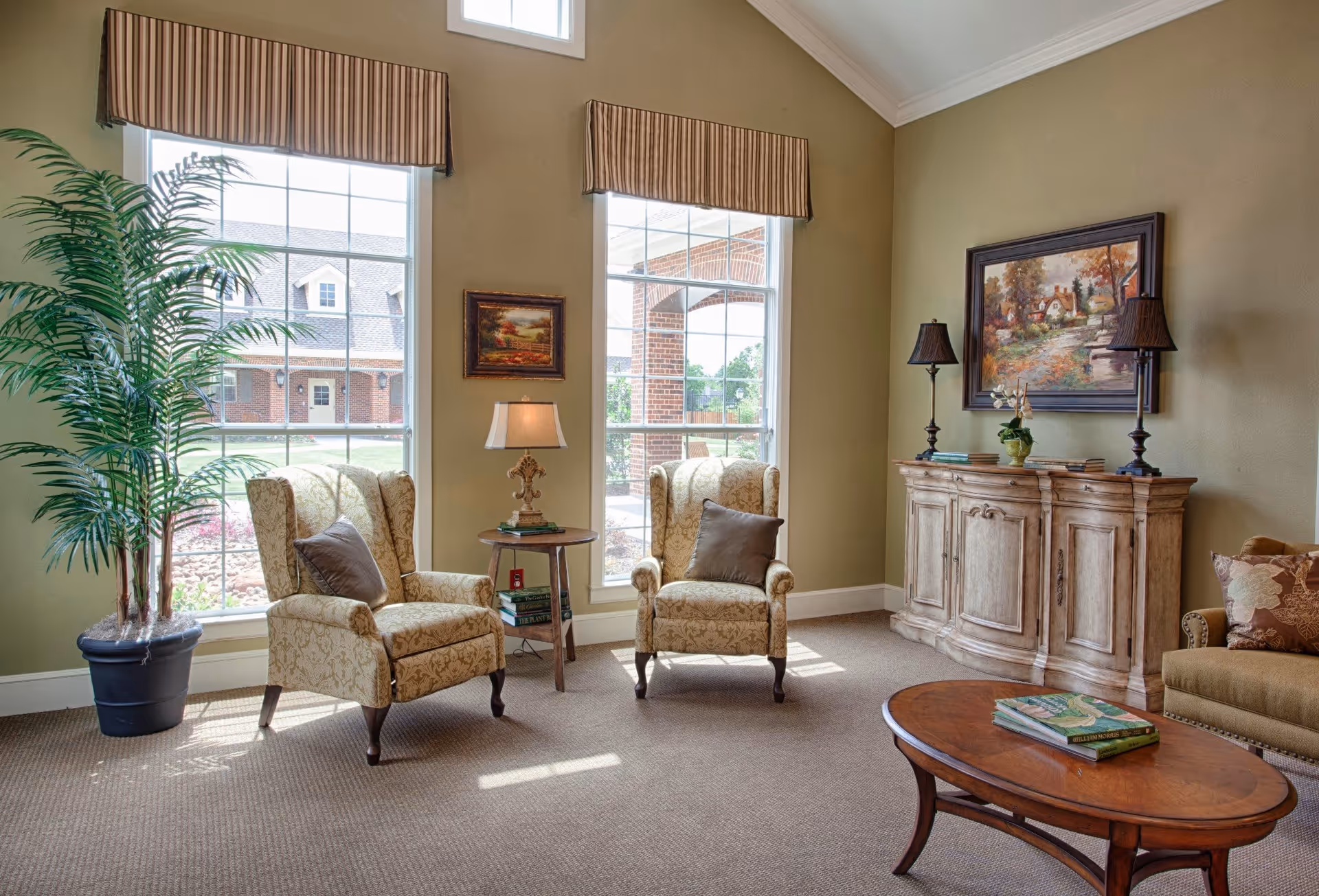 A cozy living room area with two patterned armchairs with brown pillows, a small round wooden side table with a lamp and books, a large potted plant near two tall windows with striped valances, a wooden sideboard with two table lamps and a framed painting above it, and a wooden coffee table with books on it. Outside the windows, a brick building and greenery are visible.