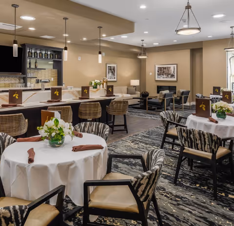A well-lit dining and lounge area in a senior living facility featuring round tables with white tablecloths, floral centerpieces, and neatly folded napkins. There is a bar counter with wicker bar stools, shelves with bottles, and a TV mounted on the wall. Comfortable seating with armchairs and sofas is arranged near a fireplace, with framed artwork on the walls and pendant lighting overhead.