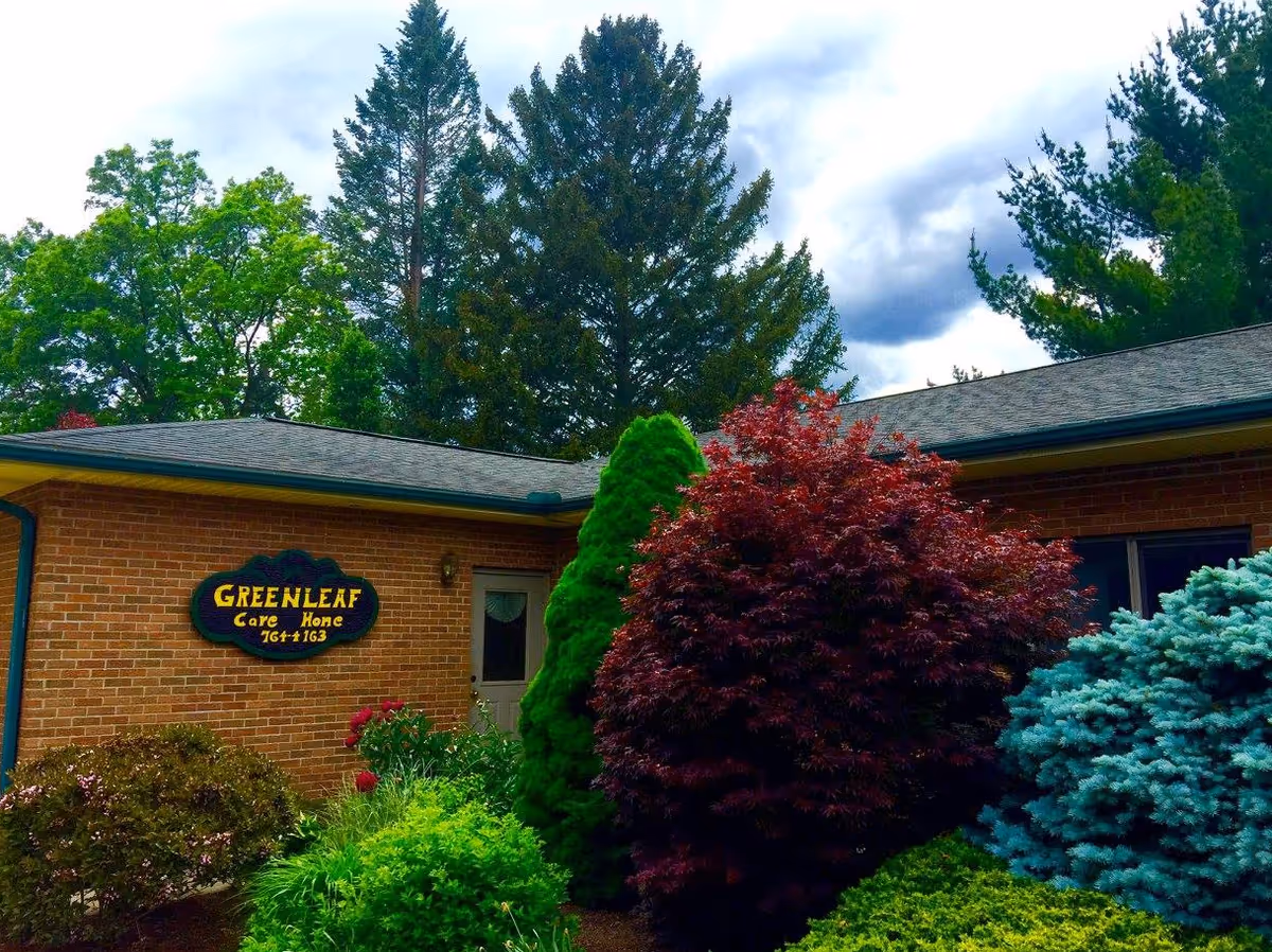 Exterior view of Greenleaf Care Home, a single-story brick building partially obscured by various green, red, and blue shrubs and trees under a cloudy sky.