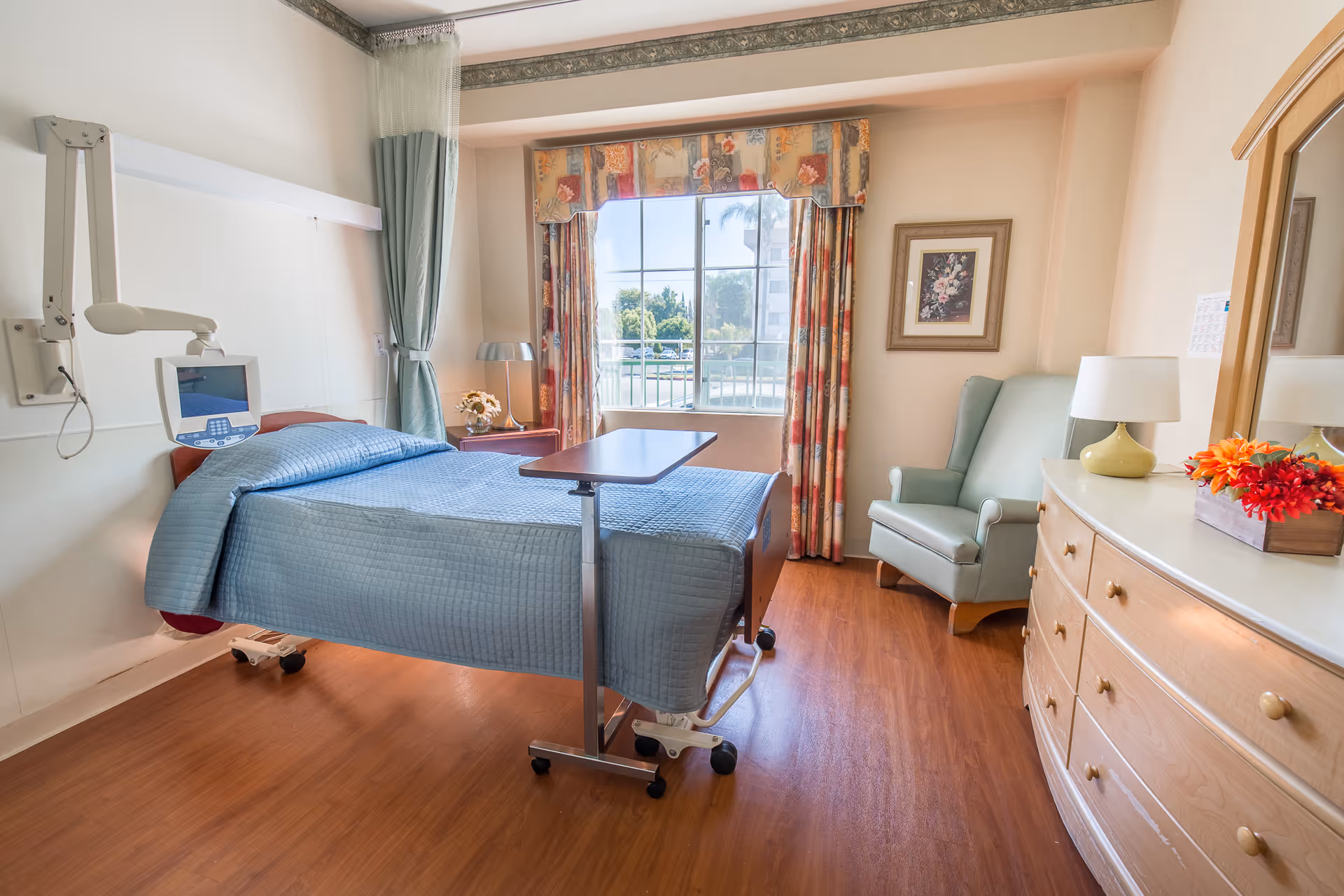 A bright and clean healthcare facility bedroom with a hospital bed covered in a blue quilt, a bedside table with a lamp and flowers, a green armchair, a wooden dresser with a lamp and a flower arrangement, and a large window with floral curtains letting in natural light.