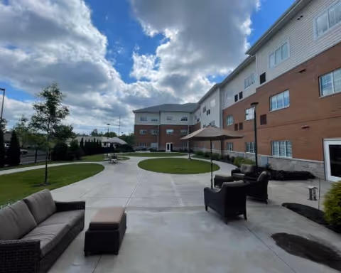 Outdoor patio area at a senior living facility with wicker seating including a sofa, chairs, and an ottoman. The patio is paved with concrete and has a large umbrella. The building is a multi-story structure with brick and siding exterior. The sky is partly cloudy.