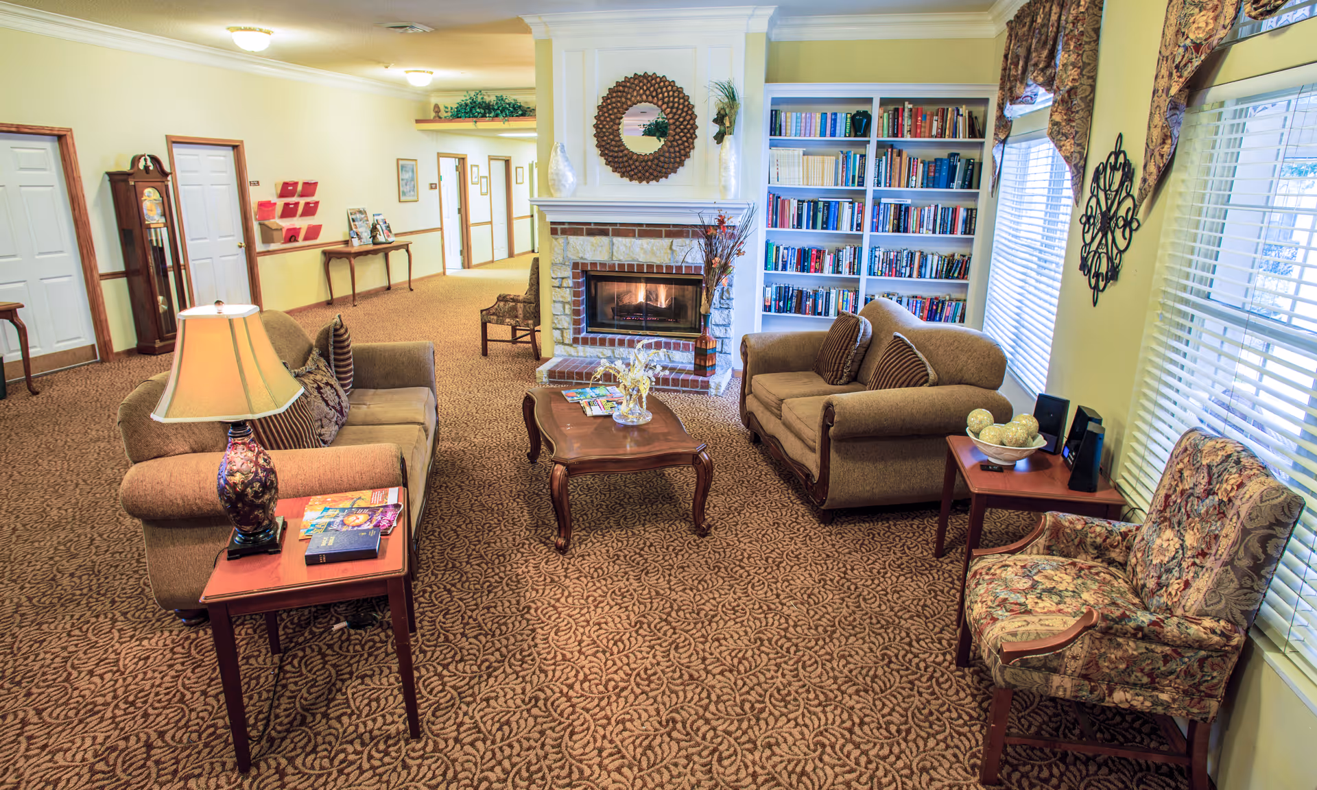 A cozy living room area in a senior living facility with two beige sofas, a floral armchair, a wooden coffee table, and side tables with lamps. There is a fireplace with a decorative mirror above it and built-in bookshelves filled with books. Large windows with floral curtains let in natural light.