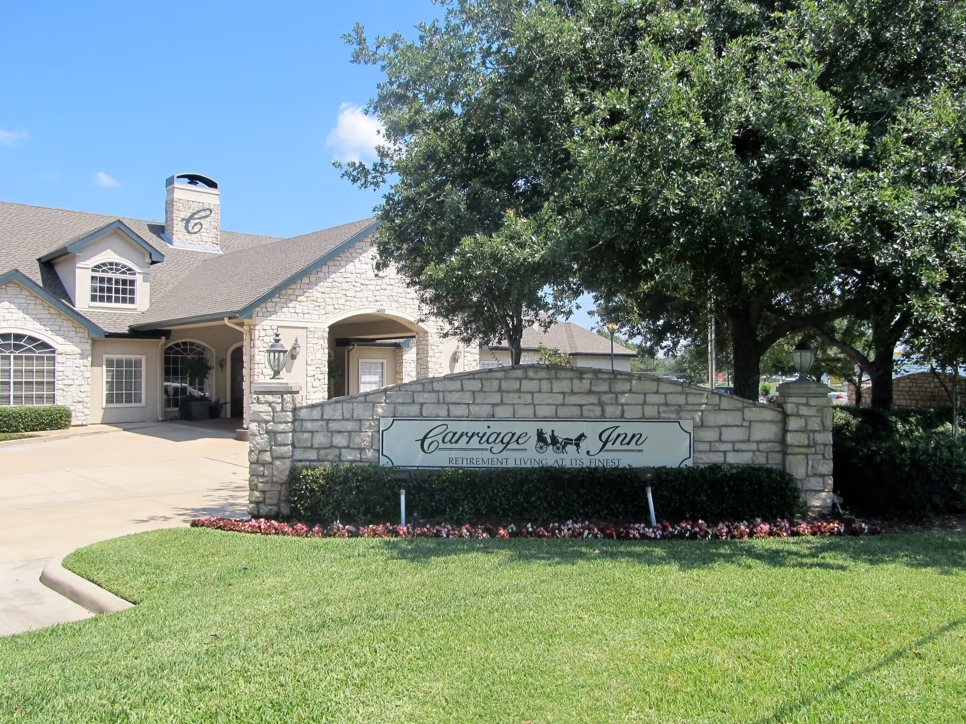 Exterior view of Carriage Inn Katy retirement living facility showing a stone building with a chimney and arched windows, a driveway, a stone sign with the facility name, and well-maintained landscaping including green grass, bushes, flowers, and trees under a clear blue sky.