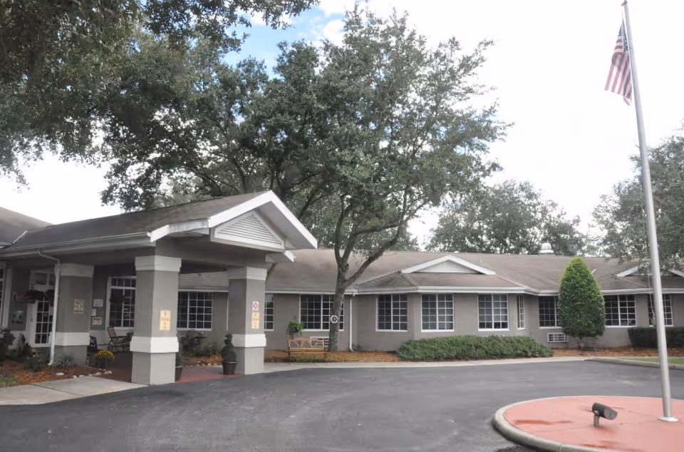 Exterior view of Crown Pointe Senior Living facility showing a single-story building with multiple windows, a covered entrance supported by columns, trees, shrubs, and an American flag on a flagpole in front.