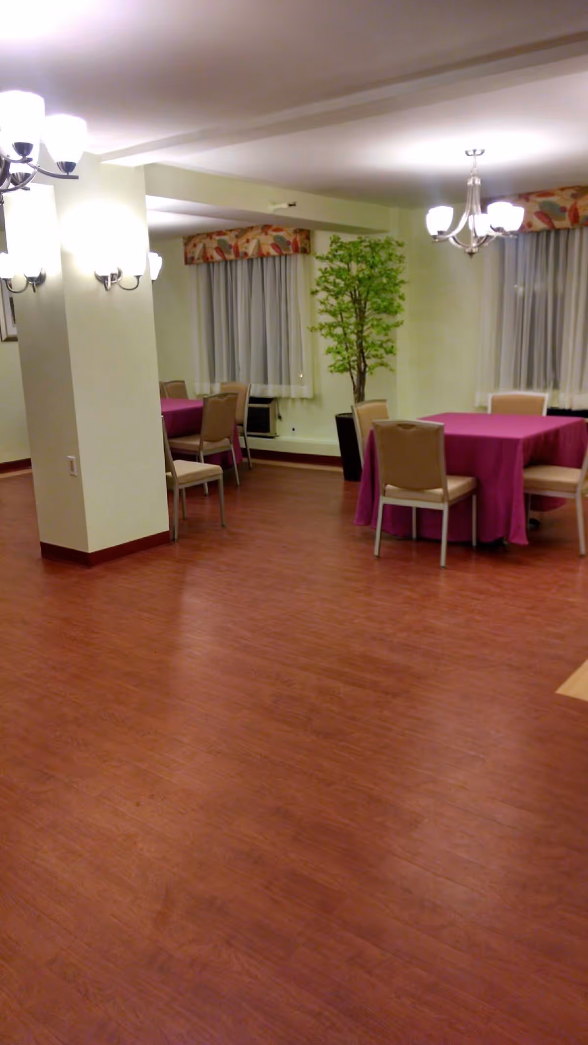 Interior view of a senior living facility dining room with wooden flooring, tables covered with purple tablecloths, beige chairs, wall-mounted lights, chandeliers, large windows with curtains, and a potted green plant.