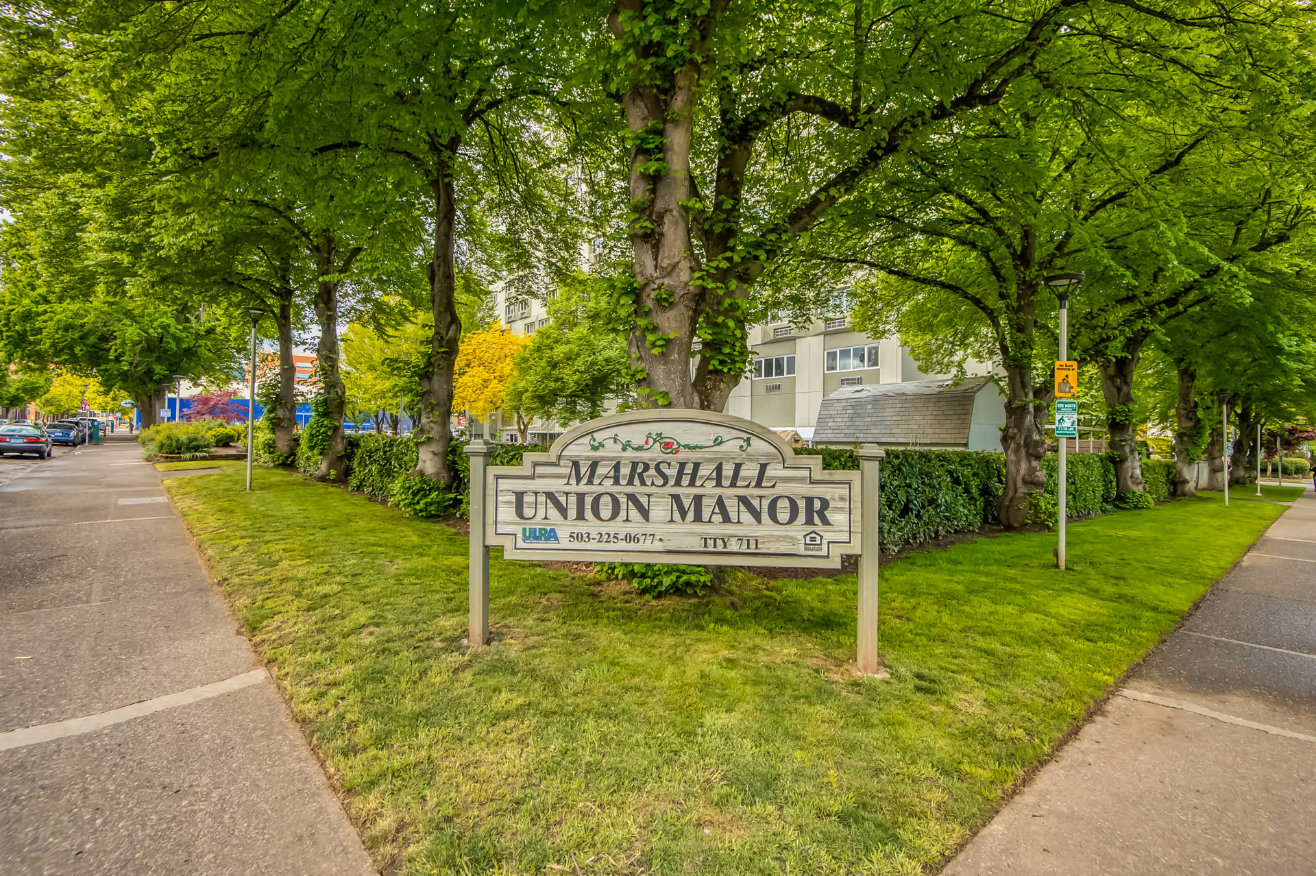 A wooden sign for Marshall Union Manor is displayed on a grassy area between two sidewalks, surrounded by green trees and bushes. In the background, there is a multi-story building partially visible behind the trees. Cars are parked along the street on the left side.