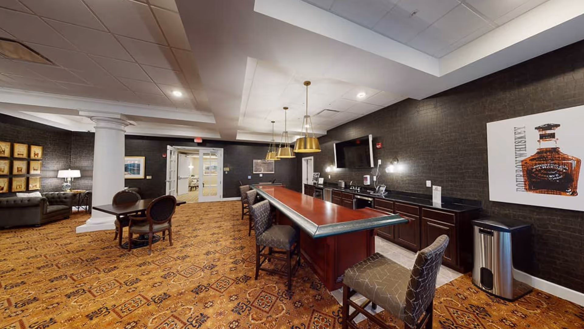 Interior view of a senior living facility lounge area featuring a long wooden bar with several cushioned bar stools, pendant lights hanging from the ceiling, dark textured walls, a mounted flat-screen TV, and a framed artwork of a bourbon whiskey bottle. To the left, there is a small table with chairs and a seating area with two gray tufted sofas and a lamp on a side table. The floor is covered with a patterned carpet.
