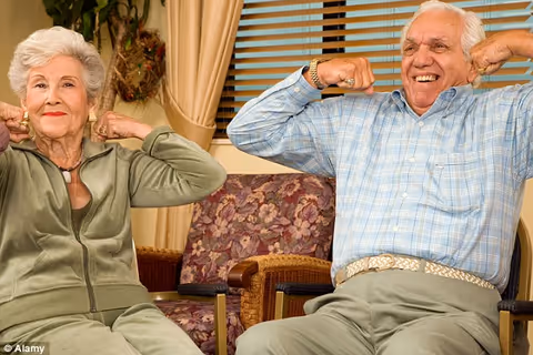 An elderly woman and an elderly man sitting on chairs indoors, both smiling and stretching their arms behind their heads as if exercising or relaxing. The room has floral patterned chairs, beige curtains, and a window with horizontal blinds.