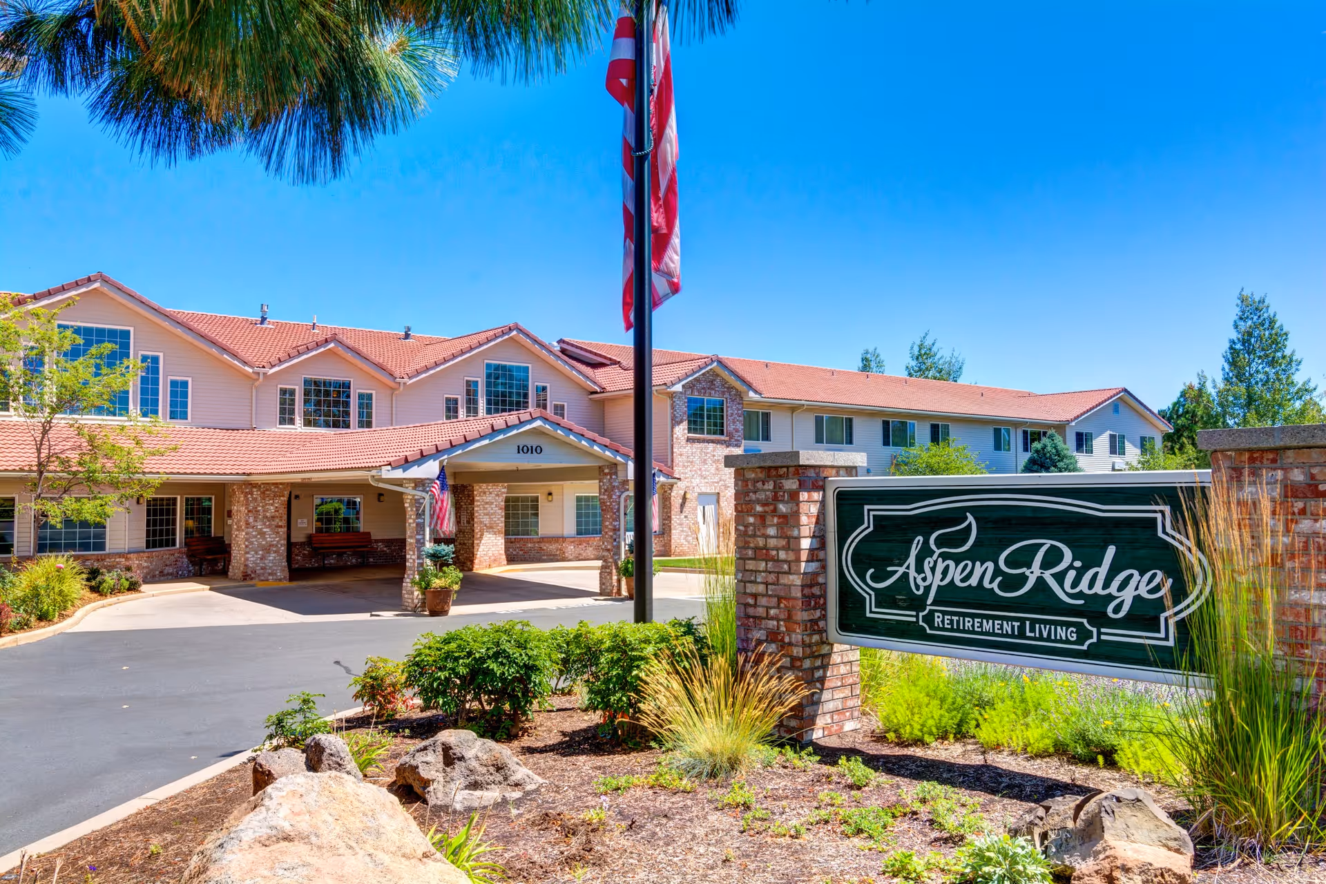 Exterior view of Aspen Ridge Retirement Community building with a covered entrance, brick and siding facade, and a sign in the foreground displaying the community name. The scene includes landscaping with rocks, plants, and trees under a clear blue sky.