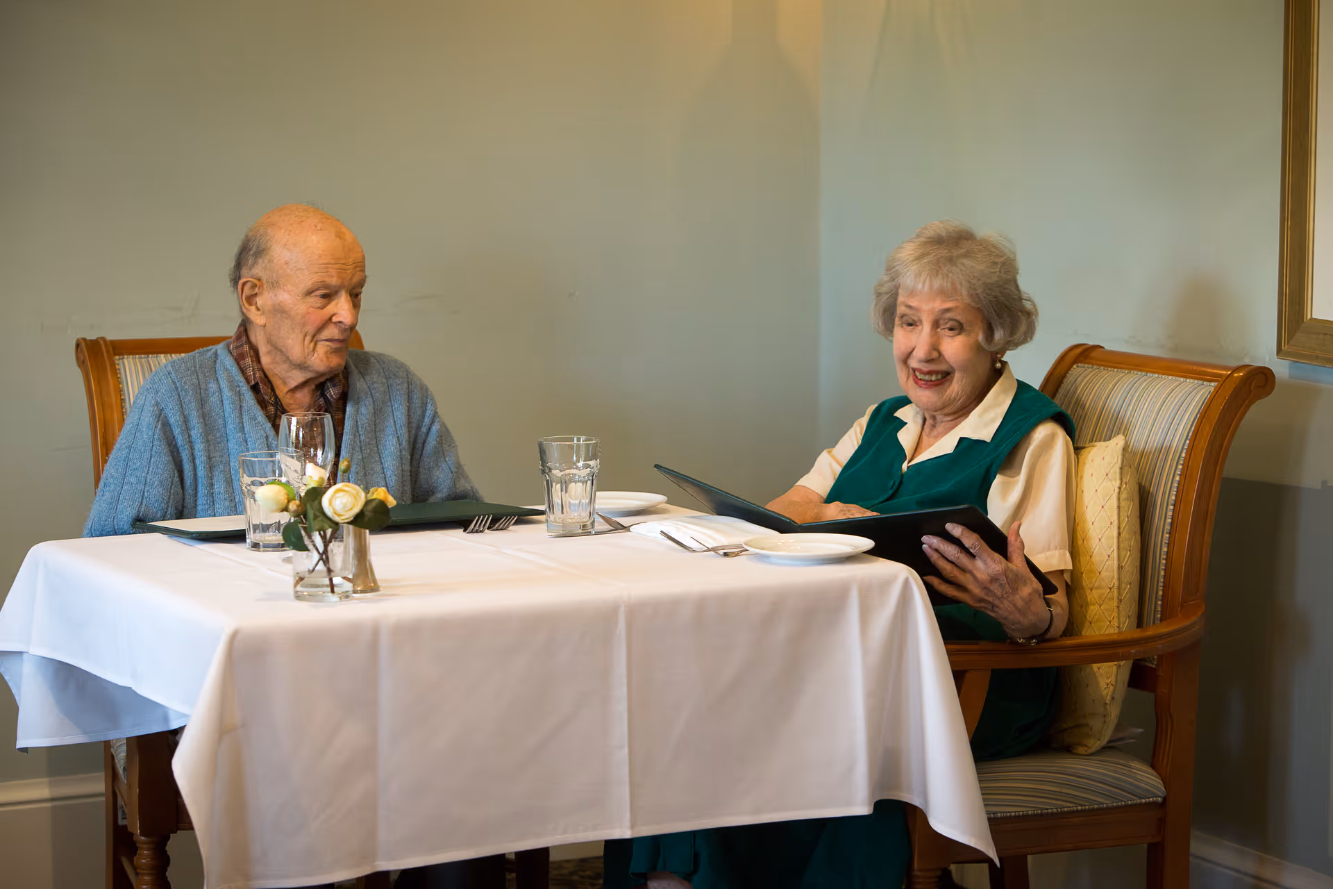 An elderly man and woman sitting at a dining table with a white tablecloth in a well-lit room. The man is wearing a blue cardigan and the woman is wearing a green dress with a white blouse underneath. The woman is holding a menu and smiling, while the man looks on. The table has glasses, plates, cutlery, and a small vase with white and yellow flowers.