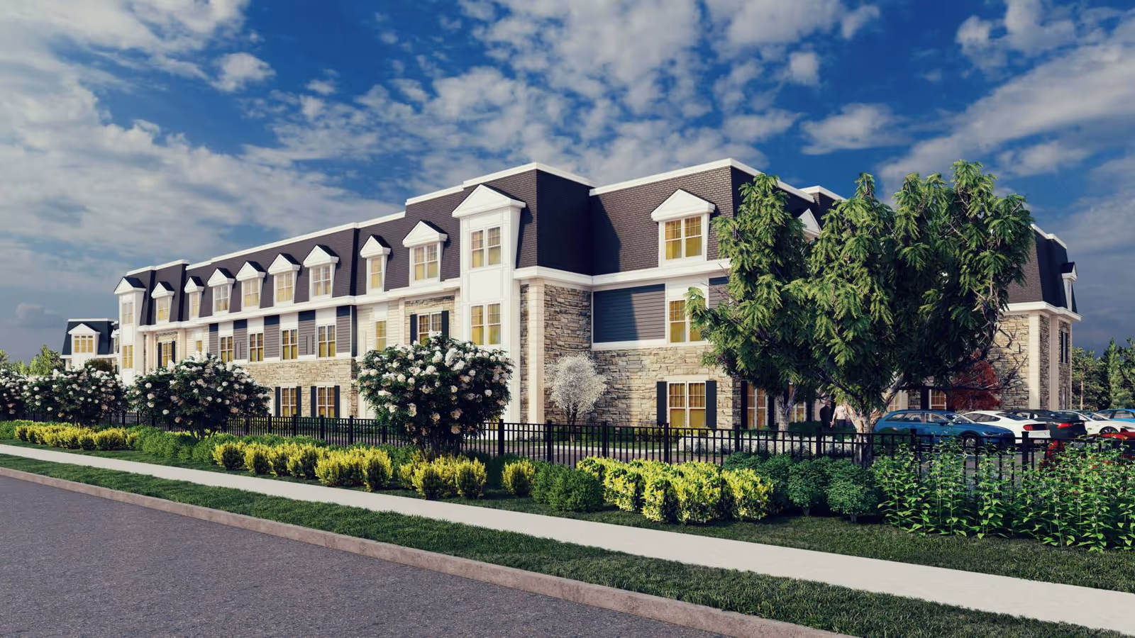 Exterior view of a large, multi-story senior living facility building with stone and siding facade, multiple windows, and a mansard roof. The building is surrounded by landscaped greenery including bushes, trees, and a sidewalk along the street under a partly cloudy blue sky.