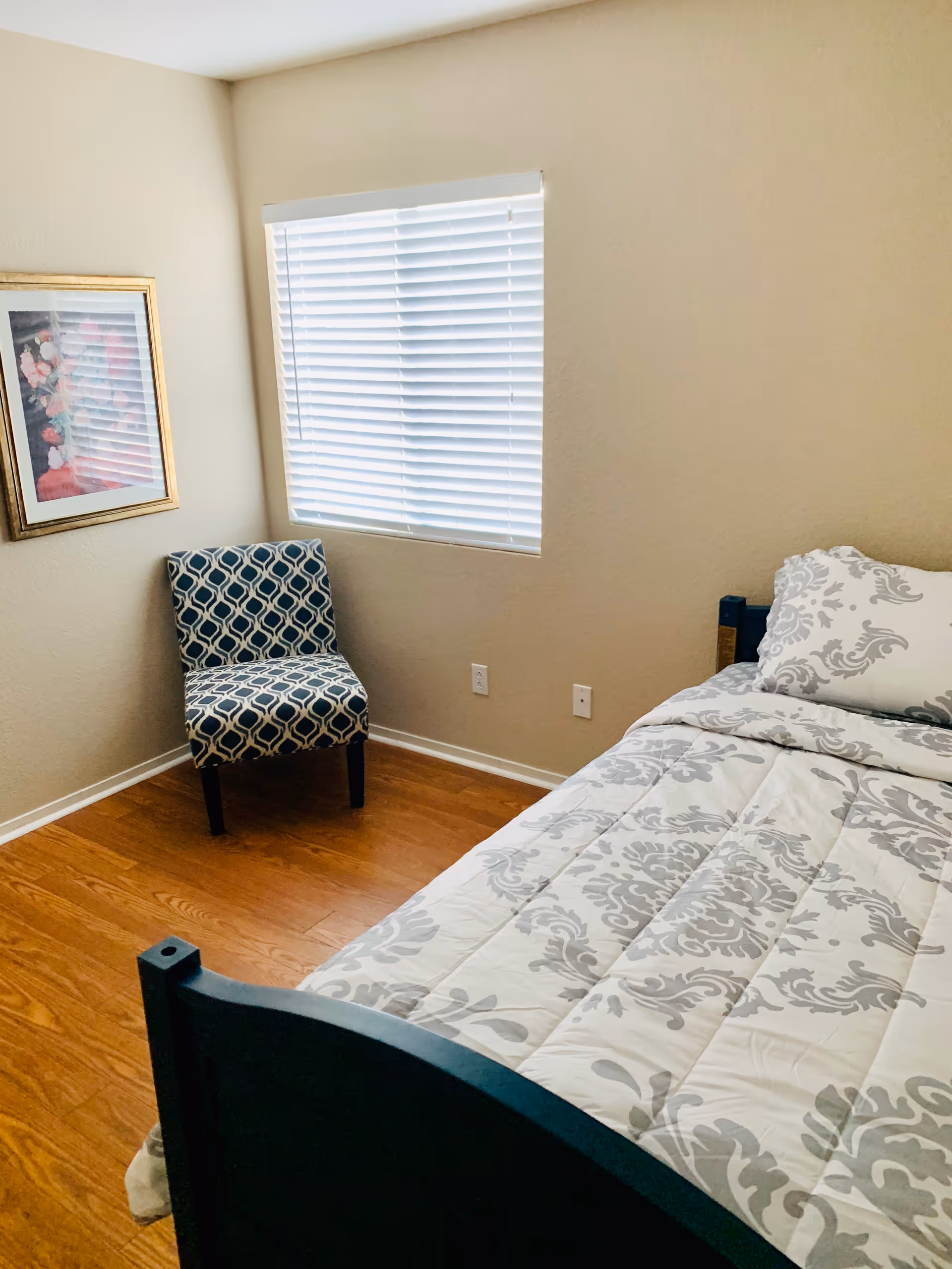 A small bedroom with a single bed featuring a white and gray patterned comforter and pillow. There is a window with white blinds on the wall, a patterned chair in the corner, and a framed floral picture hanging on the wall. The floor is wooden.