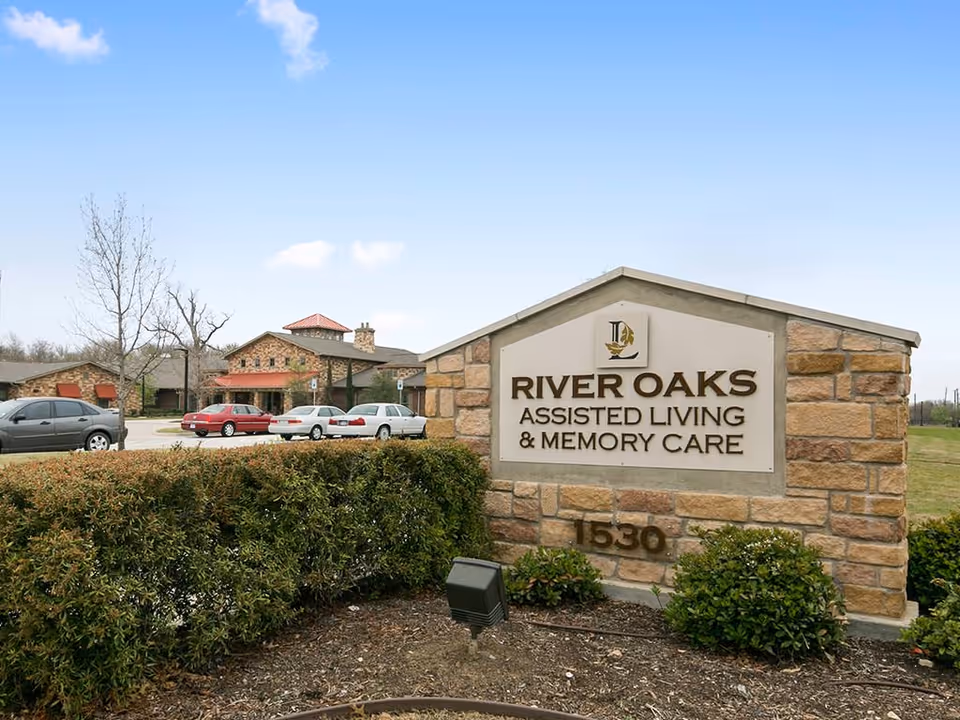 Stone sign for River Oaks Assisted Living & Memory Care with the address number 1530, surrounded by bushes and a parking lot with several cars and a building in the background under a clear blue sky.