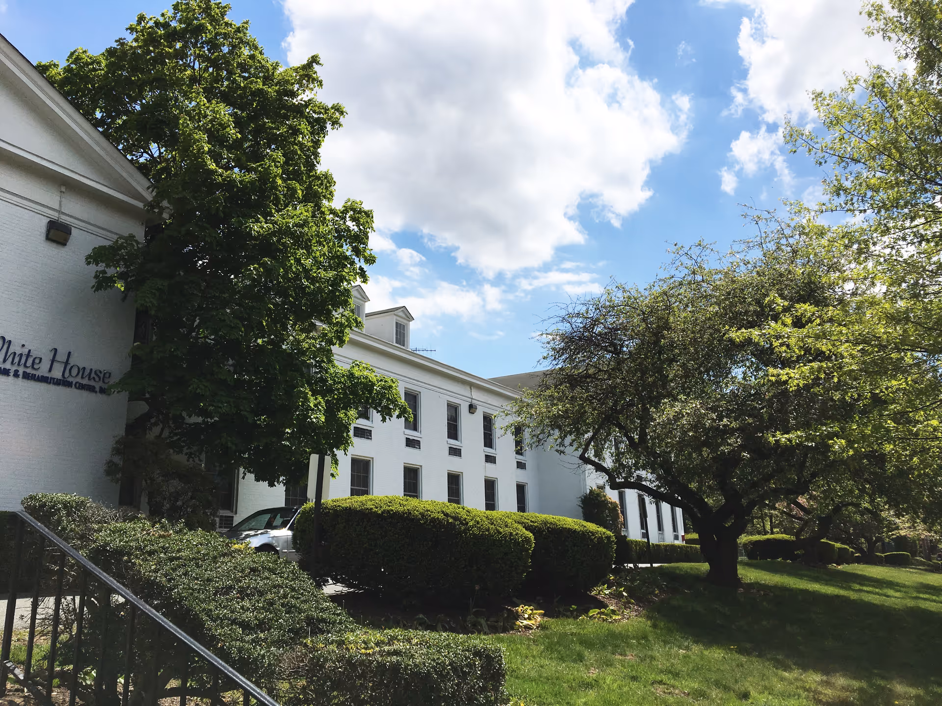 Exterior view of a white multi-story building partially obscured by green trees and bushes under a partly cloudy blue sky. The building has several windows and a sign that reads 'White House Healthcare & Rehabilitation Center'.