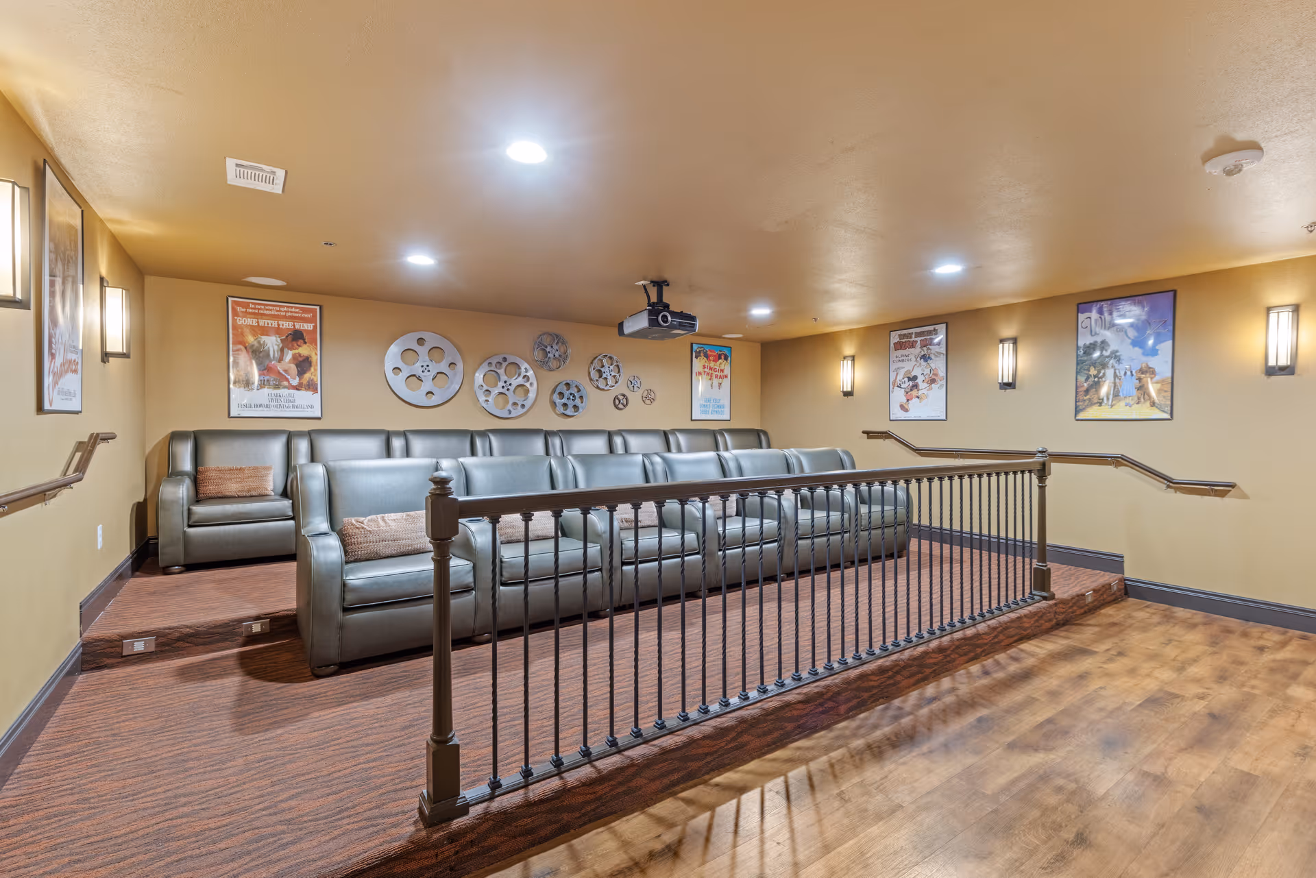A small movie theater room with two rows of green leather seats, movie posters on the walls, and decorative film reel wall art. The room has warm lighting and a projector mounted on the ceiling.