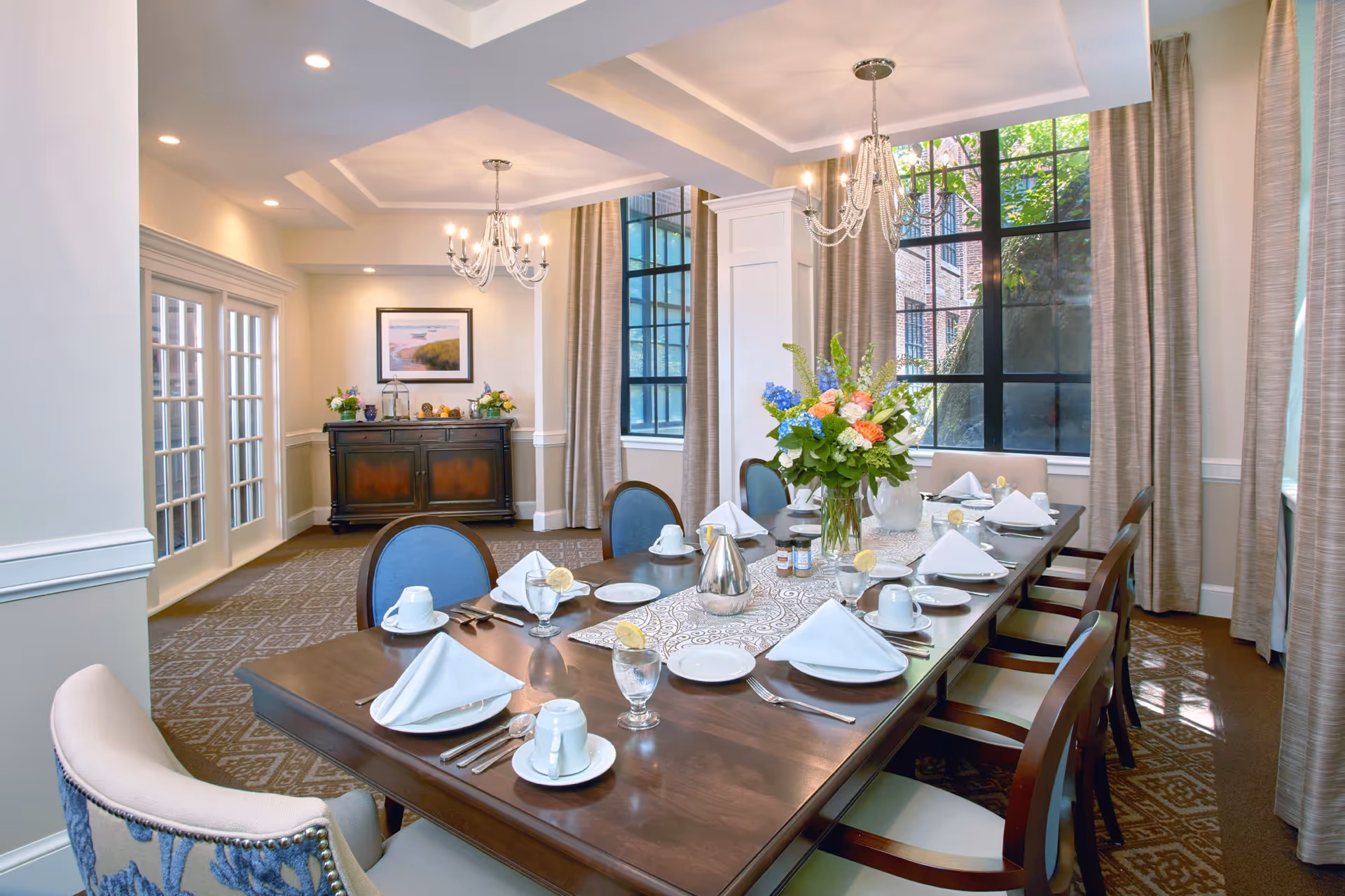 A well-lit dining room with a long wooden table set for eight people. The table is elegantly arranged with white napkins, plates, cups, glasses with lemon slices, and silverware. A floral centerpiece with colorful flowers is placed in the middle of the table. The room features large windows with beige curtains, two chandeliers hanging from the ceiling, a patterned carpet, and a wooden sideboard with decorative items and a framed picture on the wall.