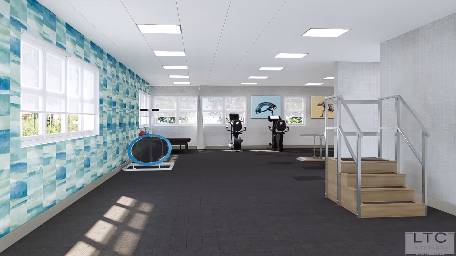 A spacious, well-lit exercise room with large windows on two walls. The room features a patterned blue and green accent wall, exercise equipment including two stationary bikes, a small trampoline, and a set of stairs with handrails. The floor is dark, and the ceiling has multiple rectangular lights. There are framed pictures on the far wall.