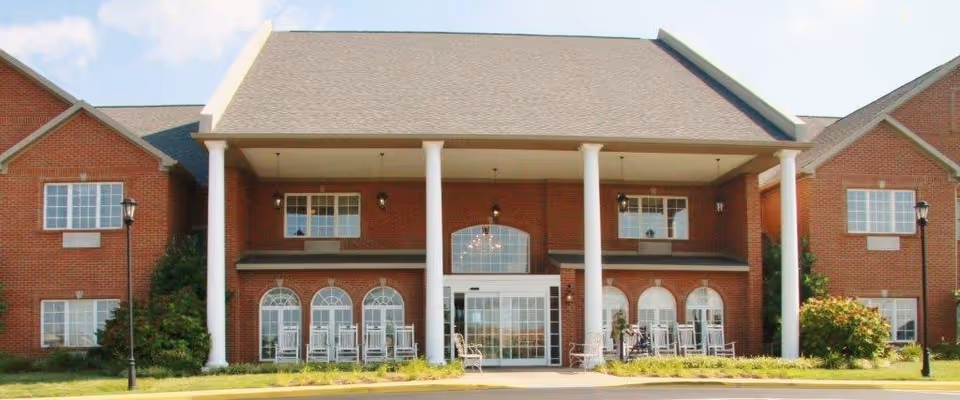 Front exterior view of a two-story brick building with white columns supporting a covered entrance. There are multiple windows, rocking chairs on the porch, and two lampposts on the lawn in front.