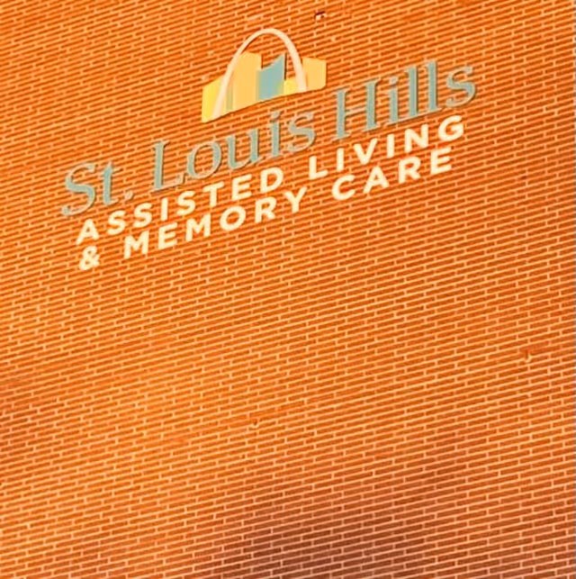 Close-up view of a brick wall with a sign that reads 'St. Louis Hills Assisted Living & Memory Care' along with a colorful arch logo above the text.