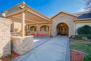 Exterior view of a senior living facility entrance with a covered pergola, stone walls, and a pathway leading to double wooden doors. There is landscaping with grass, bushes, and mulch around the building.
