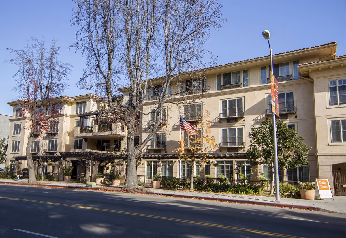Exterior view of a multi-story senior living facility building with beige walls, multiple windows, balconies, and trees in front. There is a street and sidewalk in the foreground with a streetlamp and a sign advertising daily tours.