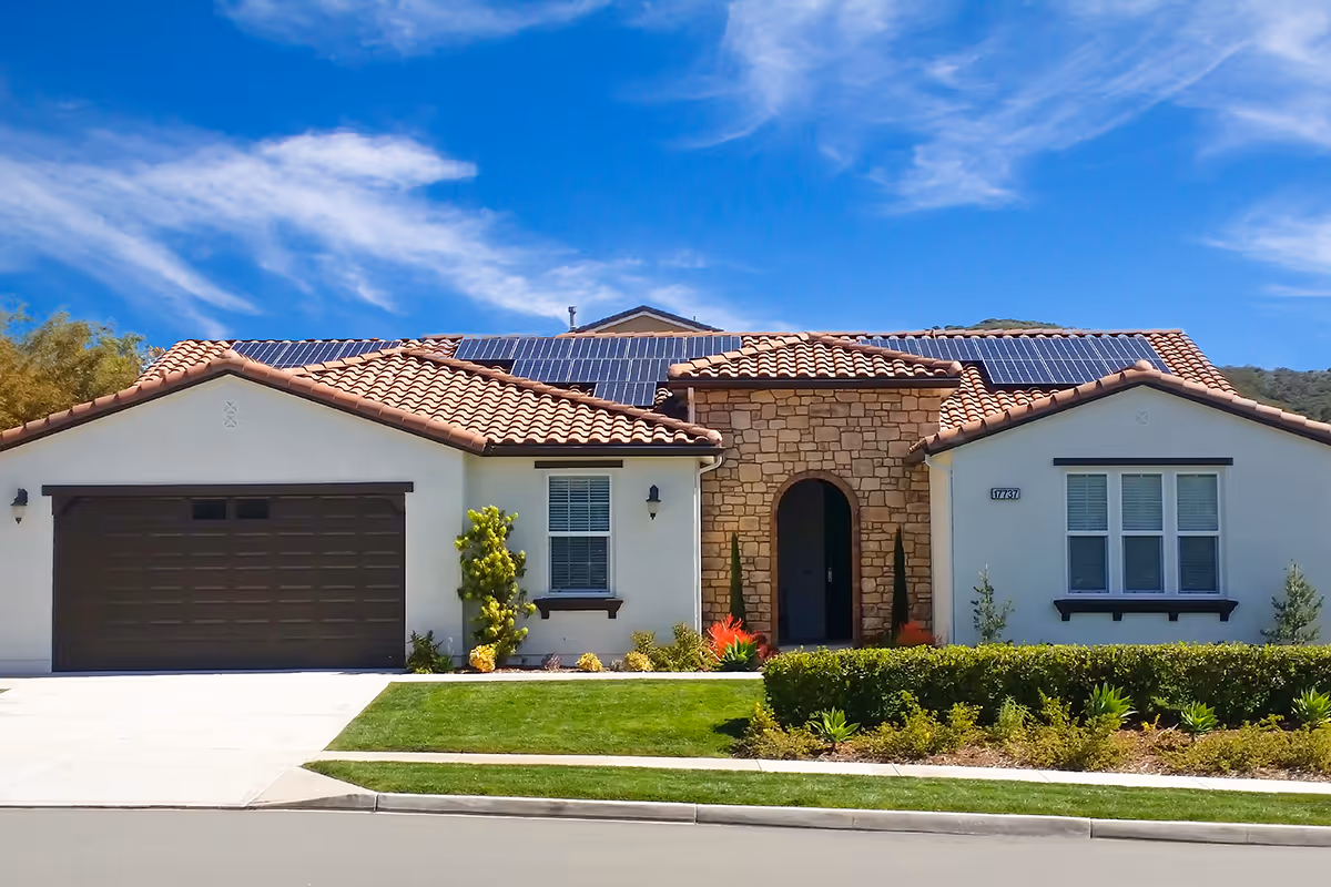 Front exterior view of a single-story house with a tiled roof and solar panels, a stone entrance, a two-car garage, and well-maintained landscaping including bushes and a green lawn under a blue sky with some clouds.