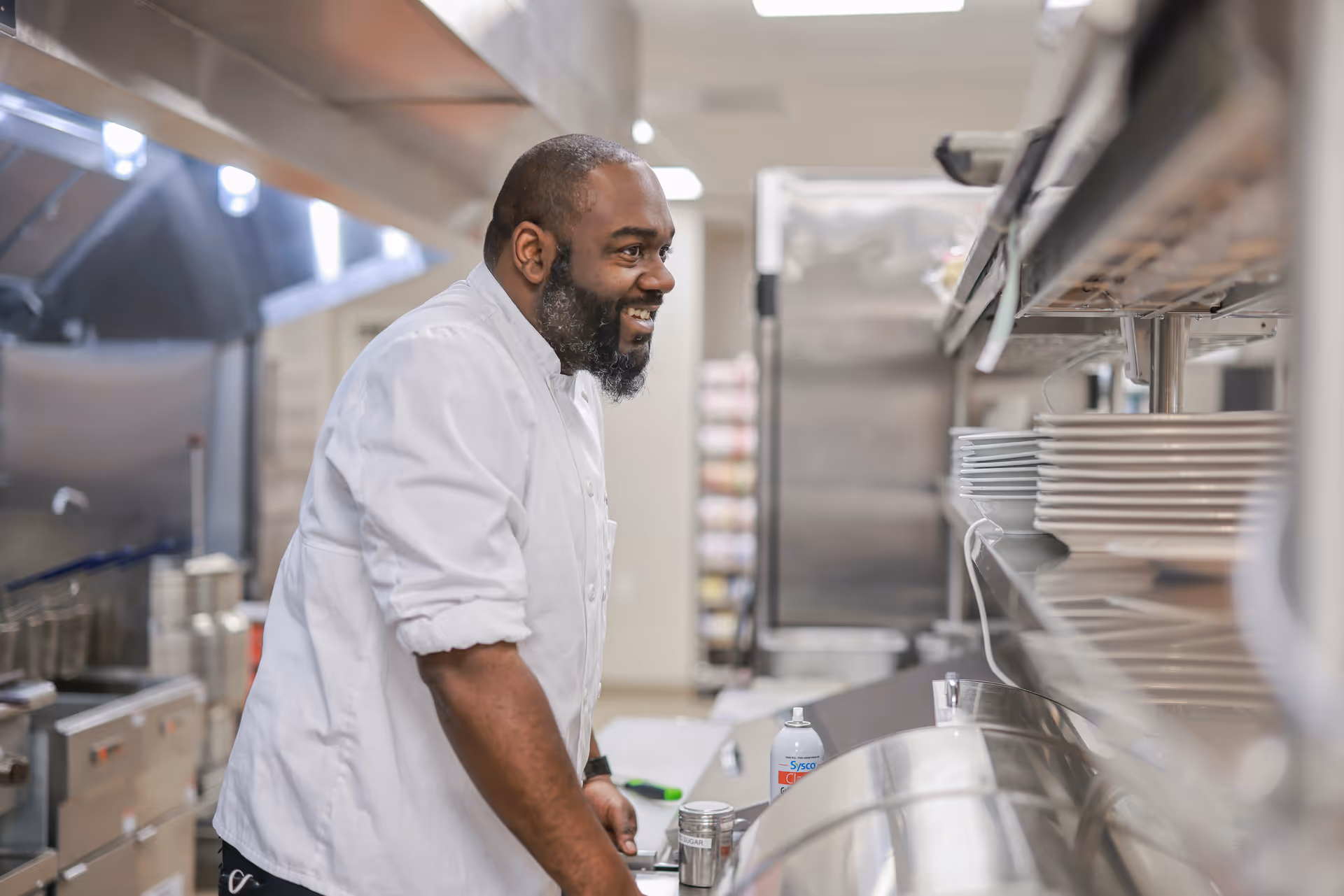 A smiling chef wearing a white coat stands in a commercial kitchen, leaning on a counter with stacked plates and kitchen equipment visible around him.
