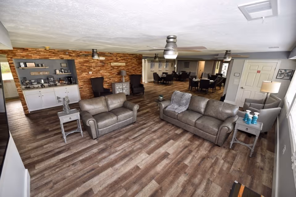 A spacious senior living facility common area with two gray leather sofas, two gray armchairs, and side tables. The room features wood flooring, ceiling fans, and a coffee station with a sign that reads 'COFFEE, ANYONE?'. In the background, there are dining tables and chairs arranged for meals. The walls are decorated with a mix of wood paneling and light gray paint.
