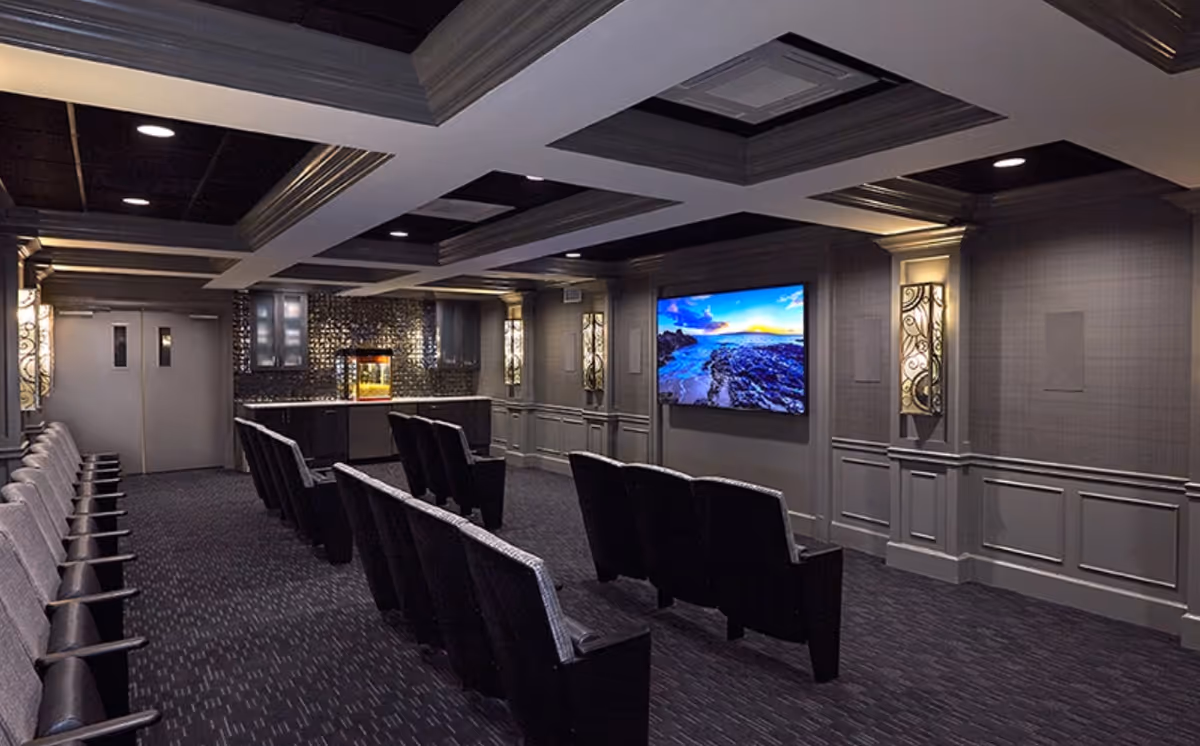 A modern theater room with rows of cushioned seats facing a large flat-screen TV mounted on the wall. The room has dark carpeting, decorative wall sconces, and a popcorn machine in the back corner near double doors.