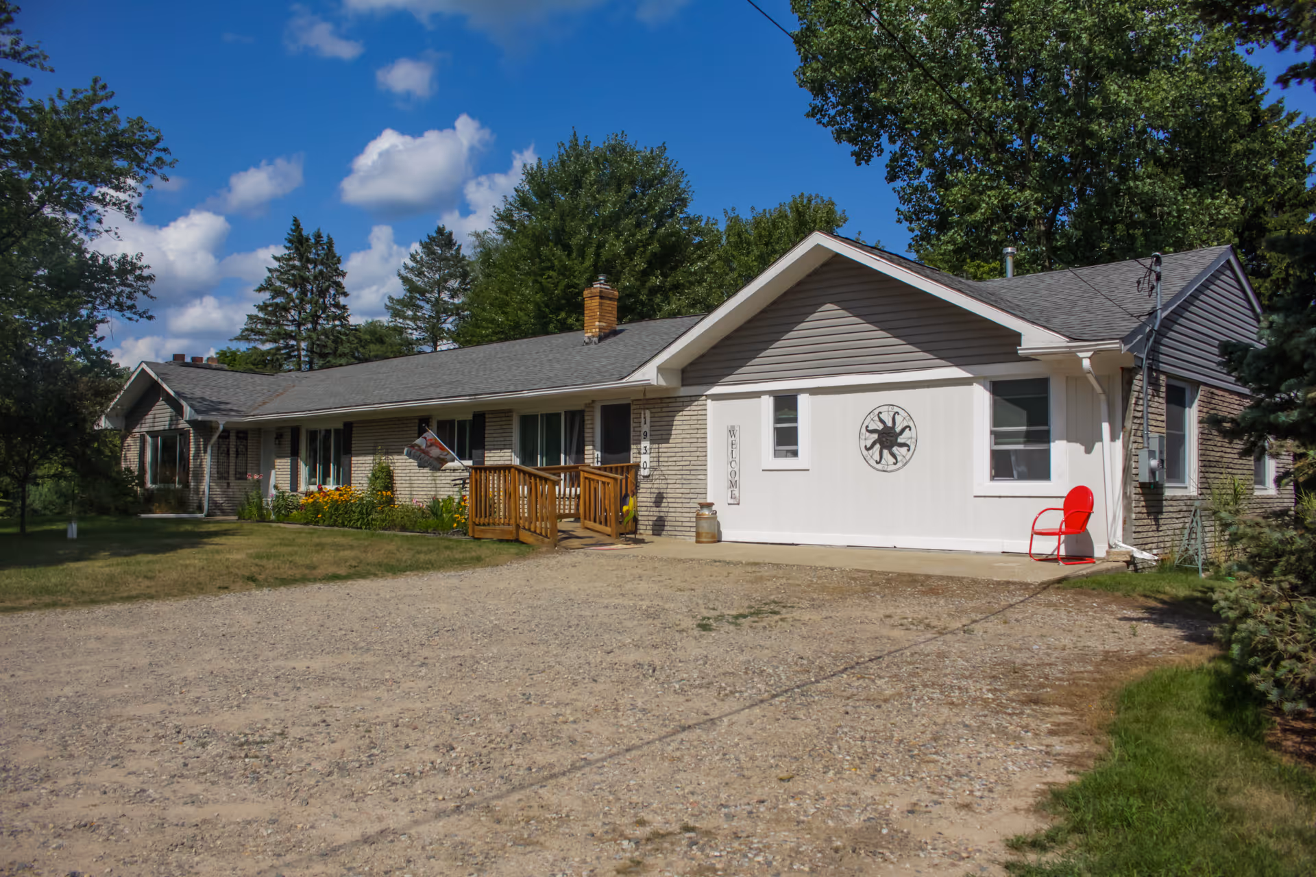 Exterior view of a single-story adult care home building with a gravel driveway, a wooden ramp leading to the entrance, a red chair on the right side, and trees surrounding the property under a blue sky with scattered clouds.