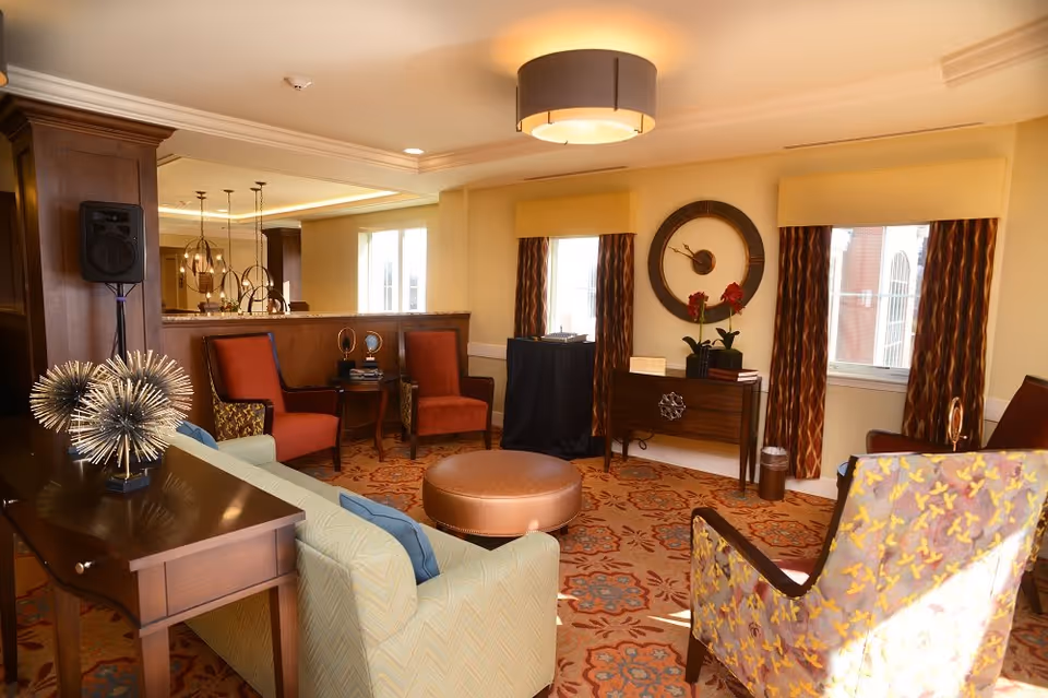 A warmly lit living room with a patterned carpet, a light green sofa with blue cushions, a round brown ottoman, two red armchairs, and a floral-patterned armchair. The room features two windows with patterned curtains, a wooden console table with a large round wall clock above it, and decorative items on the furniture.