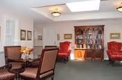 A cozy interior room with a table and four upholstered chairs in the foreground, two red armchairs against the wall, a wooden bookshelf filled with books, framed pictures on the walls, and ceiling lights with a skylight.