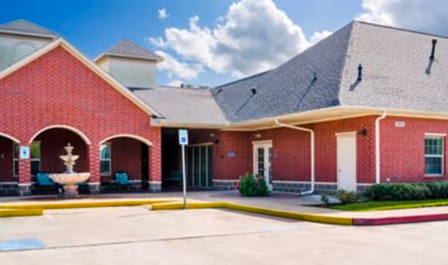 Exterior view of a red brick building with a gray shingled roof under a partly cloudy blue sky. The building has arched entryways, a fountain in front, and a parking area with handicap parking signs.