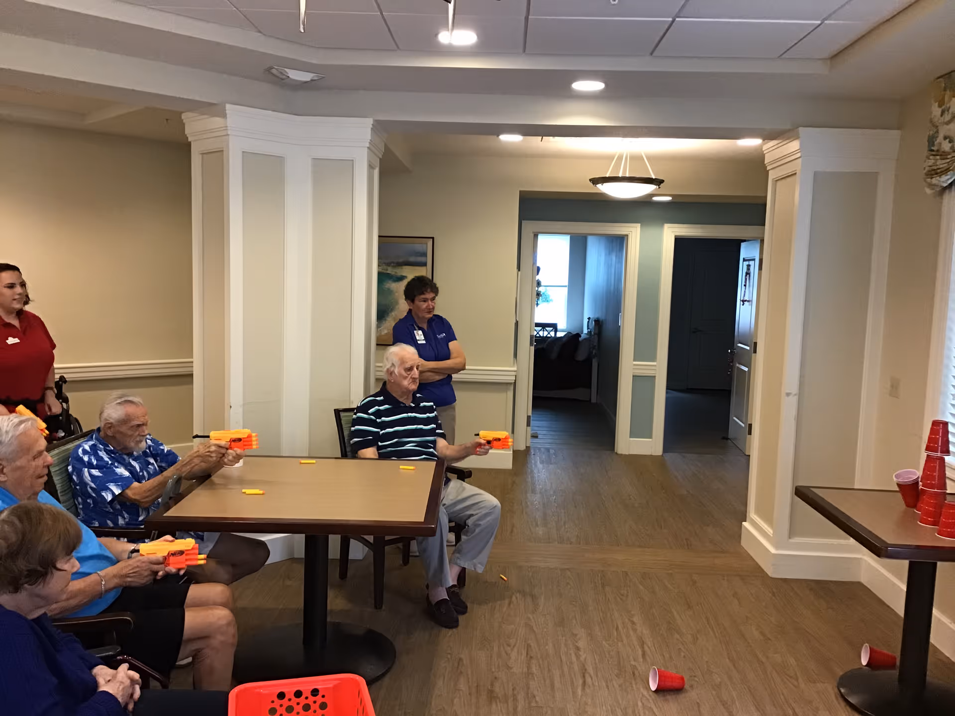 A group of elderly people sitting around a table in a common area, playing a game with toy dart guns aiming at a stack of red plastic cups on a nearby table. Two staff members stand nearby observing and assisting. The room has wood flooring, cream-colored walls, and a ceiling light fixture.