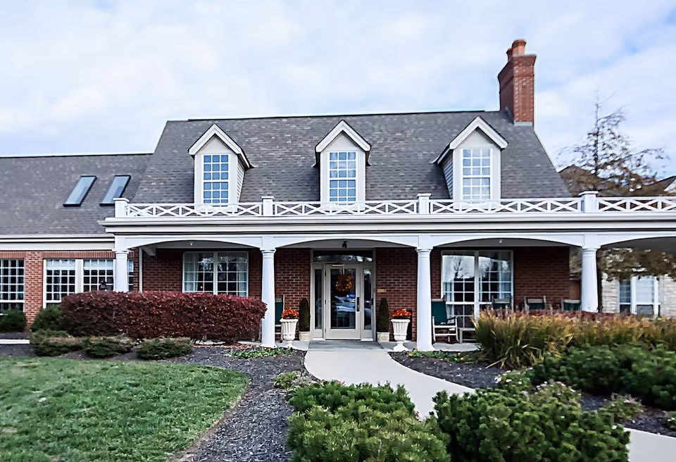 Front exterior view of a brick building with a gray shingled roof, white columns, and a covered porch with rocking chairs. There are shrubs and landscaped greenery in front of the building under a cloudy sky.