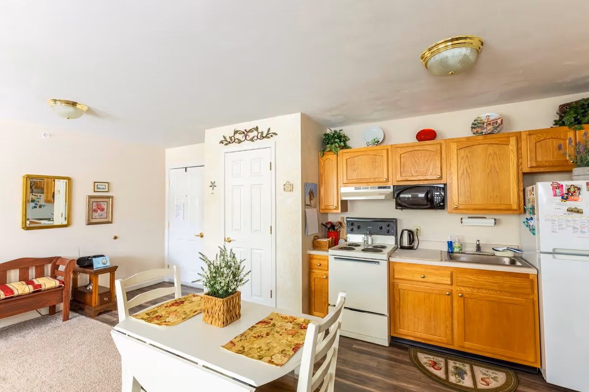 A cozy kitchen and dining area in a senior living facility featuring wooden cabinets, a white stove, microwave, refrigerator, and a sink. A small dining table with two chairs and floral placemats is in the foreground. To the left, there is a wooden bench with a cushion, a small side table with a radio, and framed pictures on the wall. The room is well-lit with ceiling lights and decorated with plants and decorative plates above the cabinets.